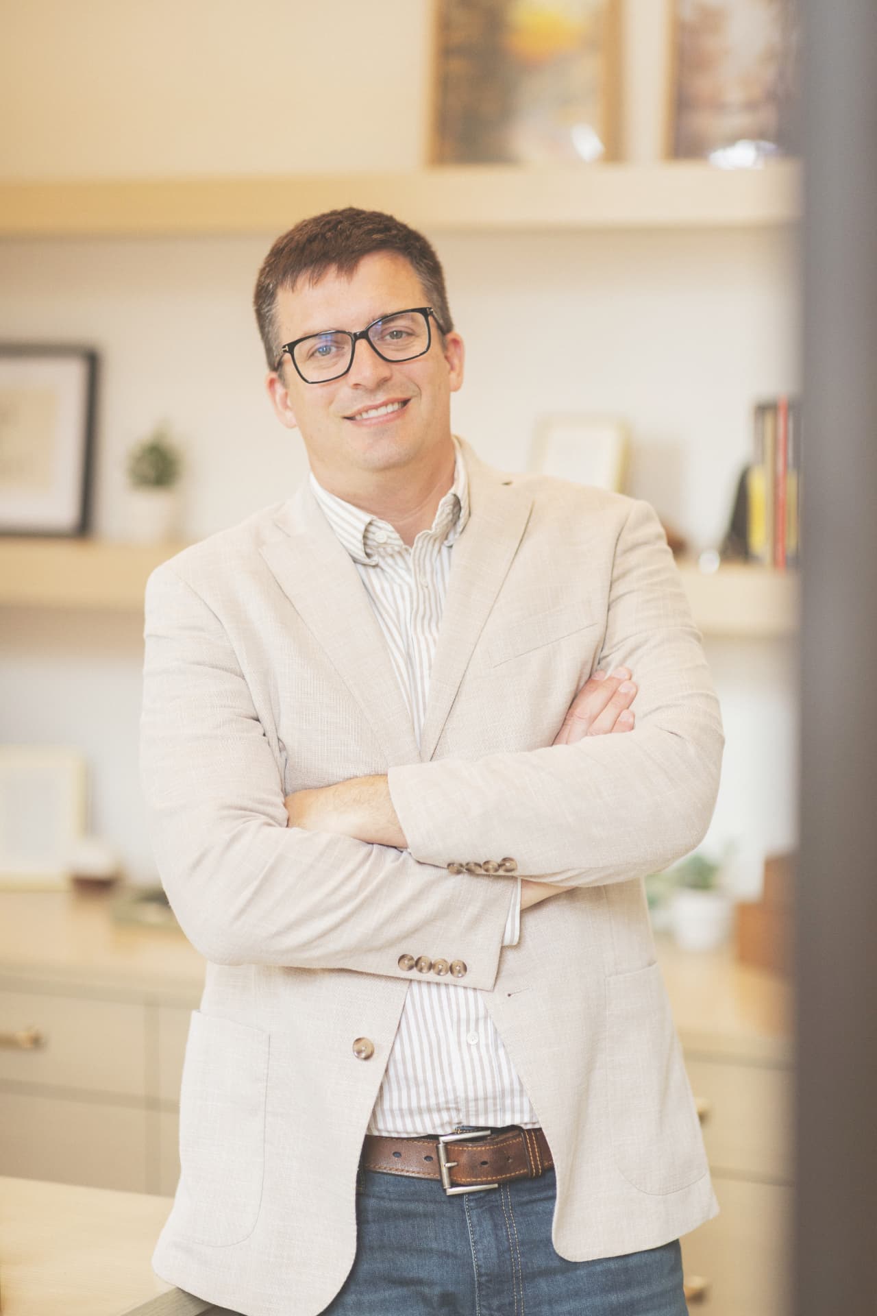 Anthony Ekren, Board Chair, smiling in a beige blazer with arms crossed in an office setting.